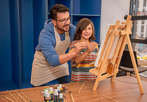 Cra 60_modelos_245 - Hombre joven pintando con un niño sobre un lienzo dentro de un salón de formación artística del Centro de bienestar integral carrera 60. 