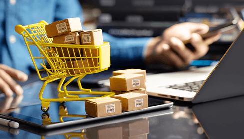  Imagen de un carrito de compras con unas cajas. Al lado se encuentra un computador portátil.