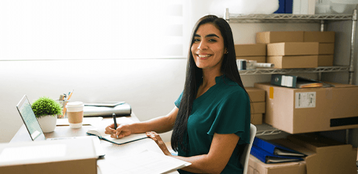 Mujer sentada sonriendo y escribiendo en una pequeña bodega de cajas.