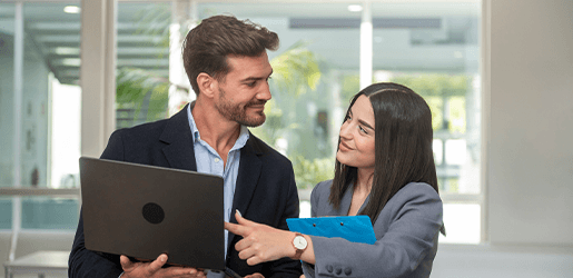 Mujer señalando la pantalla de computador, a su lado, la acompaña un hombre que sostiene un portátil.