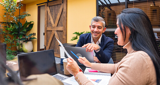 hombre y mujer sentados viendo un informe. La mujer sostiene un esfero y una hoja de papel, mientras que el hombre señala el informe.