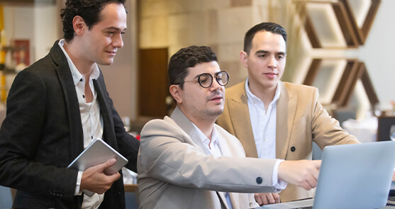 tres hombres reunidos viendo la pantalla de un computador portátil. Uno de ellos señala la pantalla, mientras que otro sostiene una tablet en su mano. 