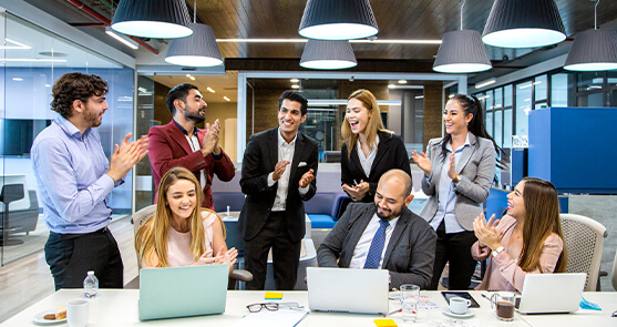grupo de oficinistas reunidos sonriendo y aplaudiendo. Algunos de ellos están sentados viendo una pantalla de computador.