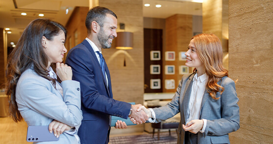 grupo de dos mujeres y un hombre con vestuario elegante. El hombre y una mujer estrechan su mano en forma de saludo. 