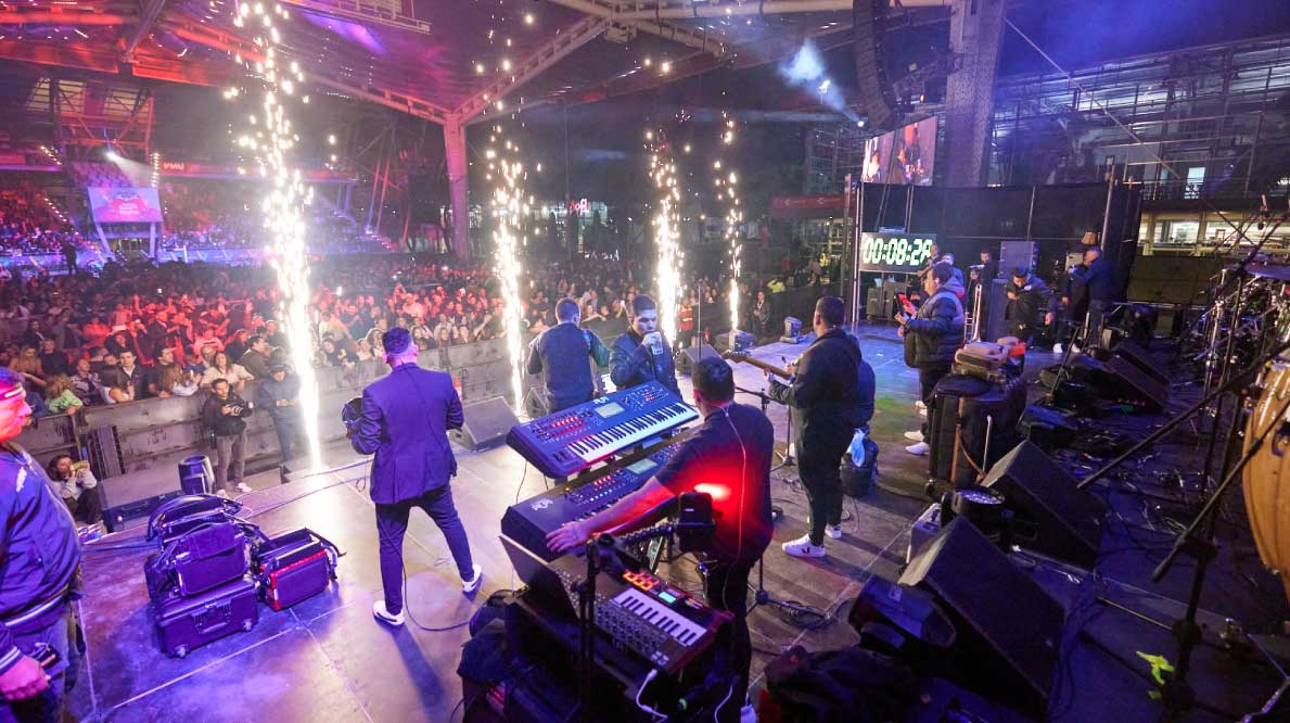 Fiesta empresarial con un escenario iluminado, música en vivo y público. 