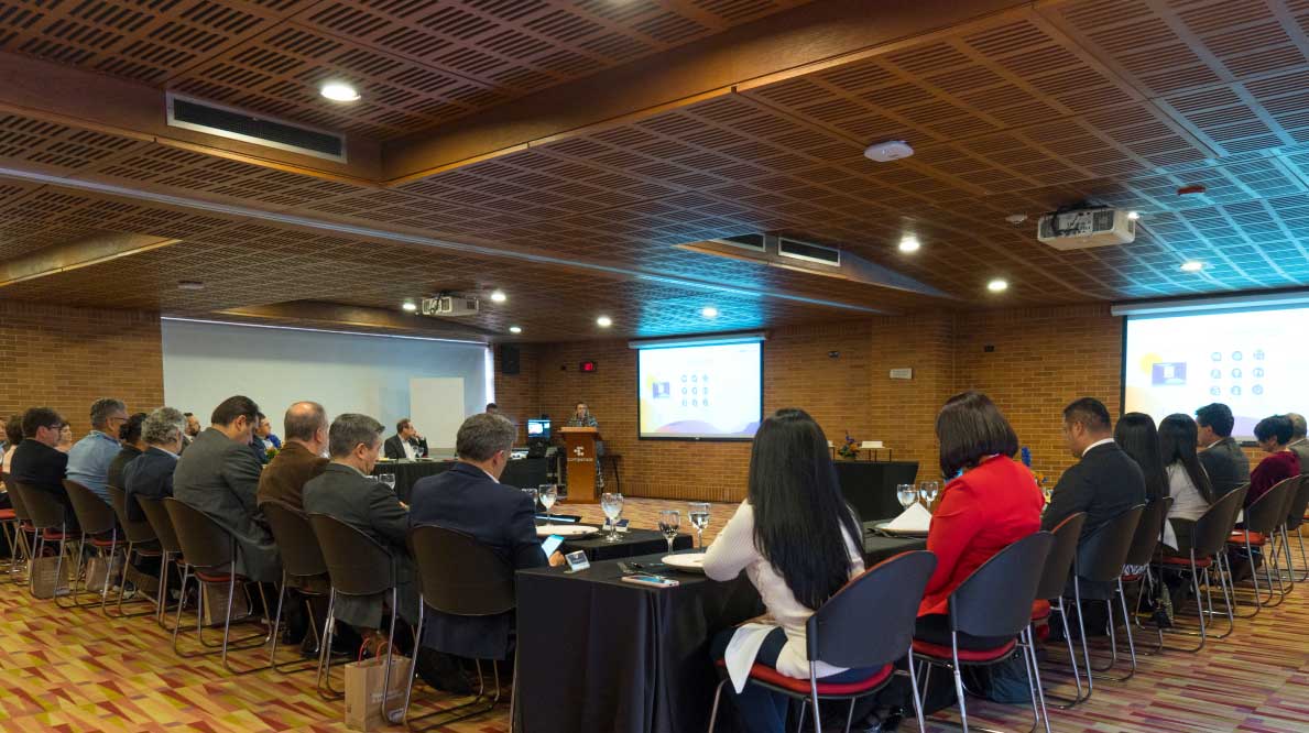  Seminario en salón con mesas redondas con asistentes y presentaciones en pantalla.