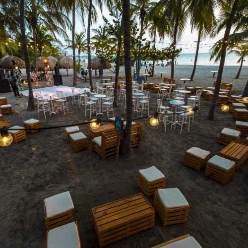 Evento en la playa con mesas y sillones de madera, rodeados de palmeras y vista al mar.