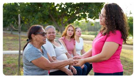  imagen de un grupo de mujeres compartiendo, dos de ellas se miran fijamente mientras se toman las manos. 