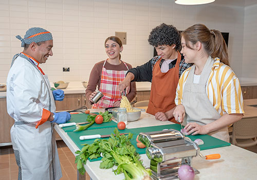 Cra 60_modelos_170 - Adultos jóvenes sonriendo con el docente de cocina, cortando vegetales y revolviendo pasta en un salón de cocina del Centro de bienestar integral carrera 60.